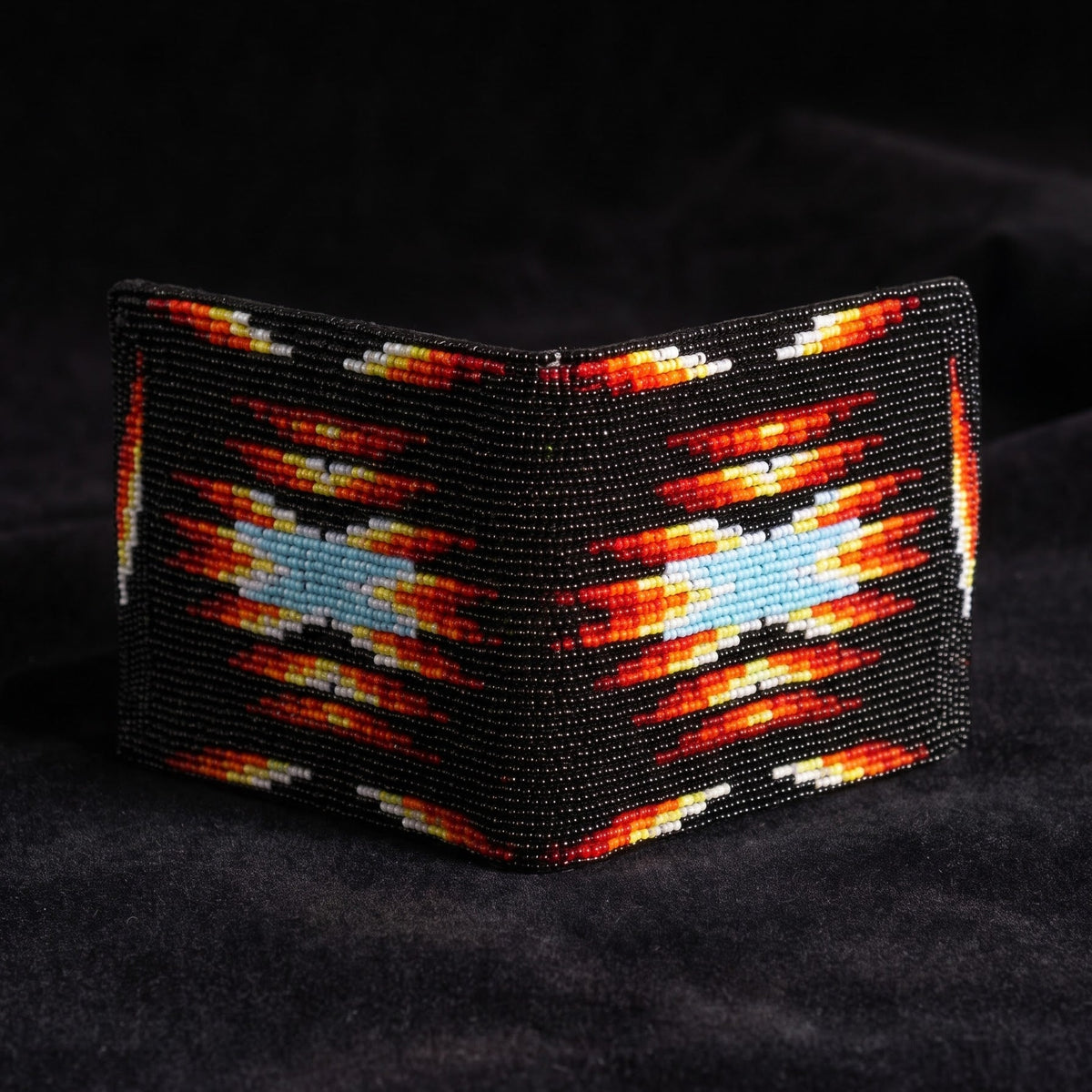A beaded wallet with a Southwestern geometric pattern in black, red, orange, yellow, and light blue, displayed on a dark textured fabric background.