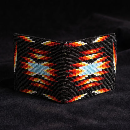 A beaded wallet with a Southwestern geometric pattern in black, red, orange, yellow, and light blue, displayed on a dark textured fabric background.