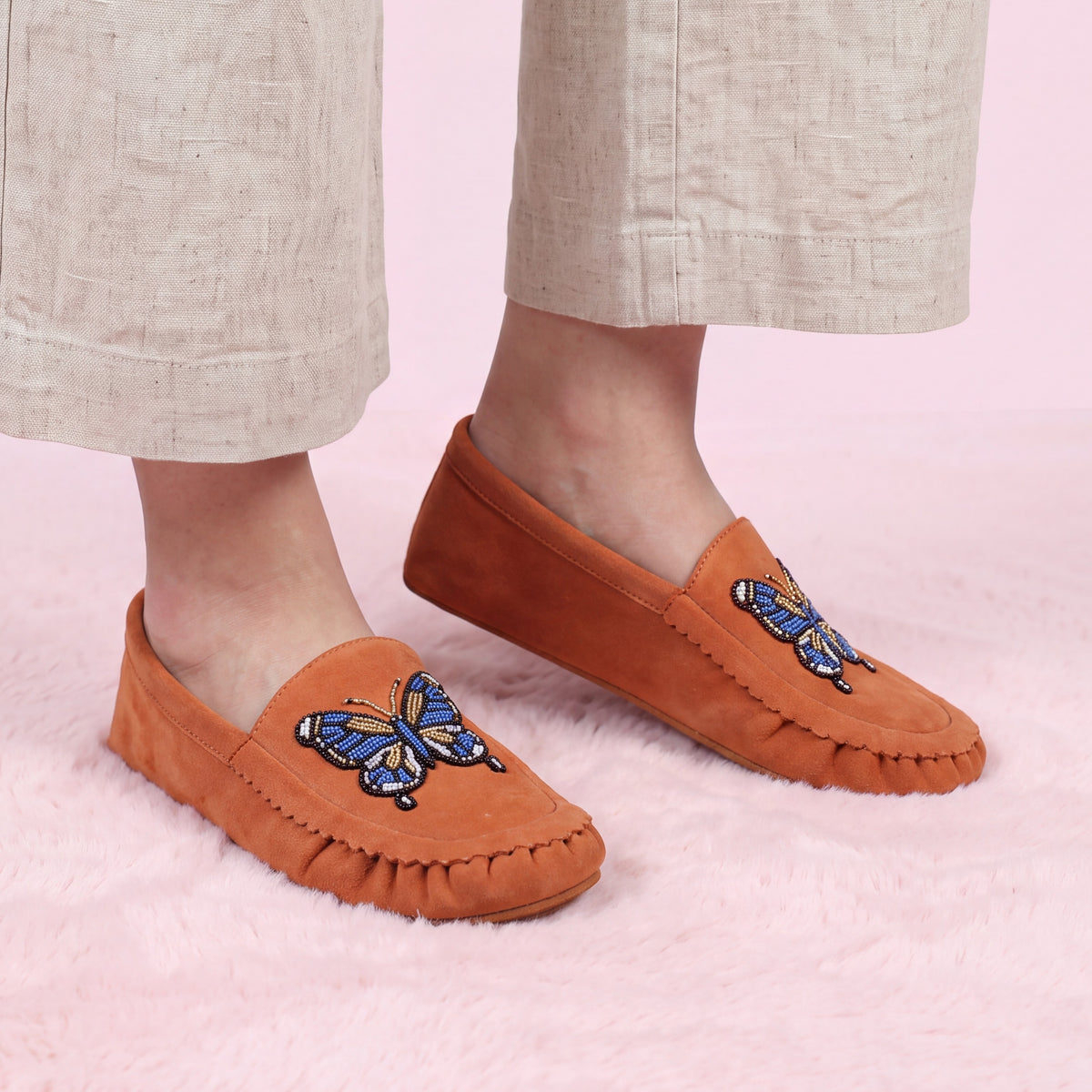 Women's feet wearing coral suede slip-on moccasins with intricate blue and gold butterfly beadwork