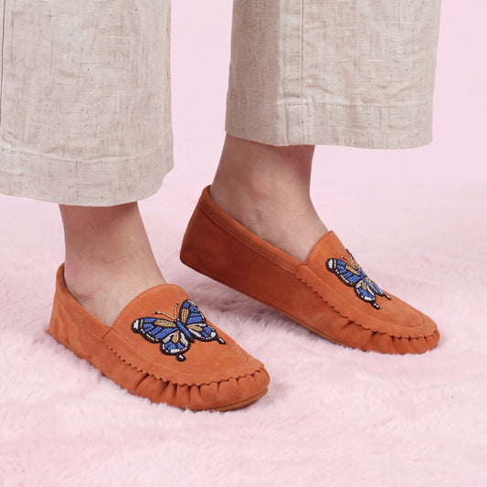 Women's feet wearing coral suede slip-on moccasins with intricate blue and gold butterfly beadwork